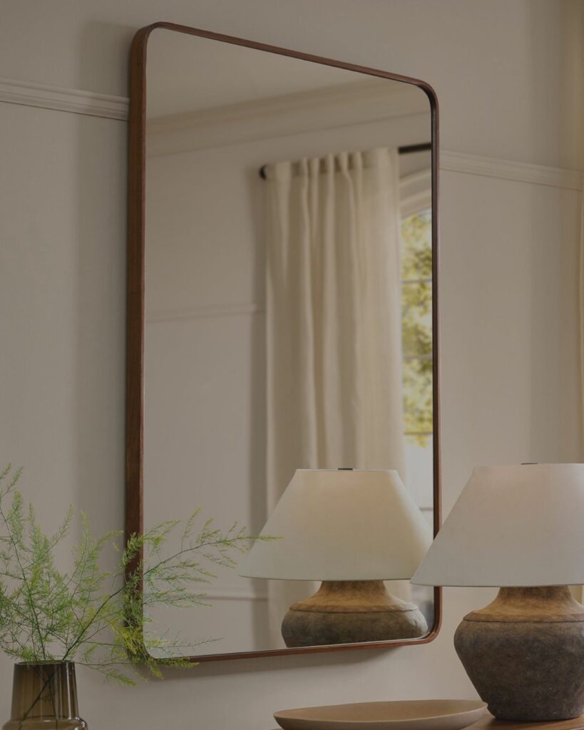 A tall, modern black arched floor mirror reflecting a bright living room with neutral linen curtains, styled alongside a ceramic table lamp on a wooden console.