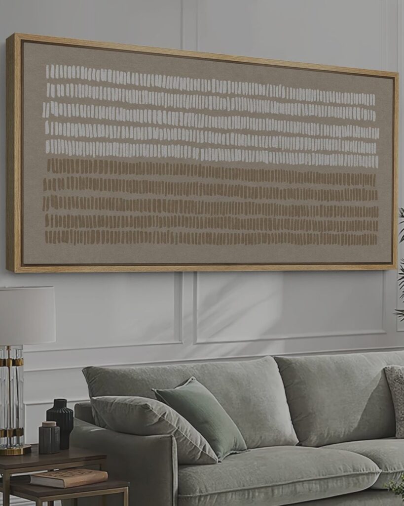 A large, minimalist oversized framed art piece hanging symmetrically above a modern neutral sofa with styled accent pillows in a bright living room.