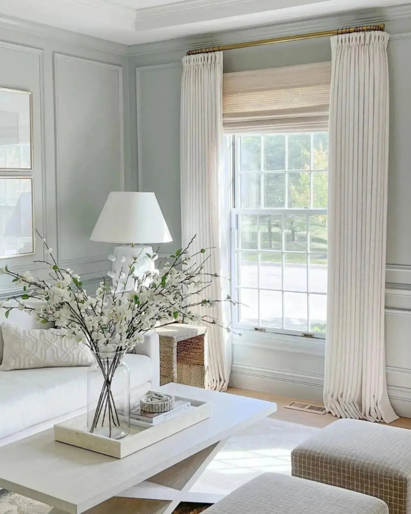 Bright luxury living room corner featuring custom cream floor-to-ceiling drapery on a brass rod paired with a woven wood Roman shade on a large window.